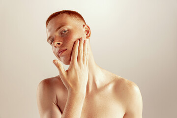 Portrait of young red-haired man with freckles touching cheeks, posing isolated over grey studio background. After shaving