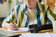 Hands of young businesswoman with arm prosthesis writing down working plan while sitting by workplace in front of laptop