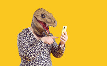 Ecentric Funny Fat Man In Dinosaur Mask Uses Mobile Applications And Communicates On Social Networks. Chubby Man With Dinosaur Head In Leopard Clothes Holding Mobile Phone On Orange Background.