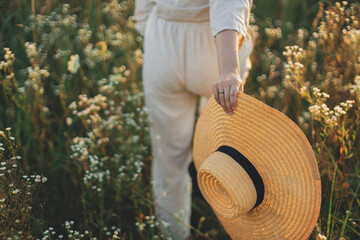 Wall Mural - Stylish boho woman with straw hat in hand close up among wildflowers in sunset light. Atmospheric moment. Summer delight and travel. Young female in rustic linen cloth walking in summer meadow
