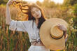 Stylish boho woman with straw hat posing among wildflowers in sunset light. Summer delight and travel. Young carefree female in rustic linen cloth relaxing in summer meadow. Atmospheric moment