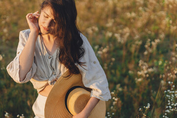 Wall Mural - Summer delight and travel. Stylish boho woman with straw hat posing among wildflowers in warm sunset light. Young female in rustic linen cloth relaxing in summer meadow. Atmospheric moment