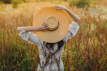 Stylish Boho Woman With Straw Hat Posing Among Wild Grasses In Sunset Light, Back View. Summer Delight And Tranquility. Young Carefree Female In Rustic Linen Cloth Relaxing In Summer Meadow