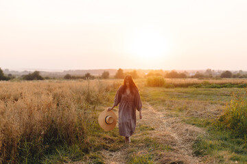 Wall Mural - Stylish woman with straw hat dancing at oat field in sunset light. Atmospheric happy moment. Young female in rustic linen dress relaxing in evening summer countryside, rural life