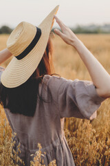 Wall Mural - Stylish woman with straw hat standing in oat field in evening light,  back view. Atmospheric tranquil moment. Young female in rustic linen dress relaxing in summer countryside. Rural slow life