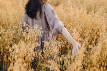 Wall Mural - Stylish woman holding oat stems in evening light, hand close up. Rural slow life. Young female in rustic linen dress standing in harvest field in summer countryside. Atmospheric tranquil moment