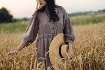 Wall Mural - Stylish woman with straw hat standing in wheat field in sunset light. Atmospheric tranquil moment. Young female in rustic linen dress relaxing in evening summer countryside. Rural slow life