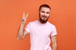 © khosrork - Portrait of friendly young adult bearded man standing with raised hand and showing v sign or peace, victory gesture, wearing pink T-shirt. Indoor studio shot isolated on orange background.