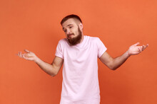 Don't Know, Who Cares. Portrait Of Bearded Man Standing With No Idea Gesture, Shrugging Shoulders Raising Hands, Wearing Pink T-shirt. Indoor Studio Shot Isolated On Orange Background.