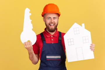 Portrait of smiling builder man holding paper house and key, looking at camera, offering help, home repair services, repair on turnkey. Indoor studio shot isolated on yellow background.