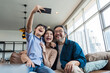 © saksit - Family smiling at camera. Happy family taking a selfie, smiling at a phone at home.