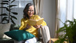 © Prathankarnpap - Happy mature woman reading book while resting on comfortable in bright living room