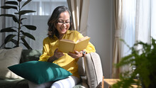 Happy Mature Woman Reading Book While Resting On Comfortable In Bright Living Room