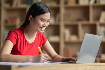 Smiling happy teenage japanese girl doing homework at table with laptop in room interior