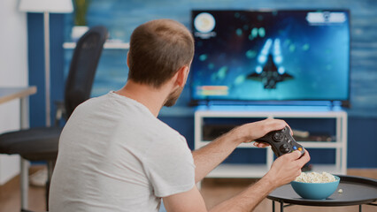 Man holding wireless controller playing console video game on tv while sitting on sofa in modern living room. Gamer relaxing on couch enjoying online gaming first person shooter on television.