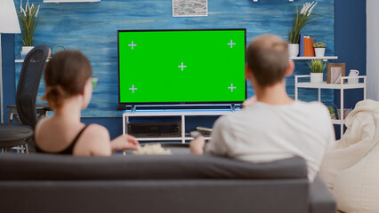 Wall Mural - Couple sharing a bowl of popcorn while watching tv and switching channels looking at green screen in modern living room. Woman and man watching chroma key television and zapping while having a snack.