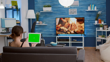 Wall Mural - Back view of young woman holding digital tablet with green screen watching social media video content sitting on sofa. Girl looking at touchscreen device with chroma key looking at influencer vlog.