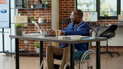 Paralyzed employee in wheelchair using laptop to plan project for startup business. Suffering from chronic impairment and physical health condition in disability friendly office. Tripod shot.