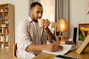 Poster - Young African American man sitting at desk, working on laptop, taking notes during webinar, drinking coffee at home