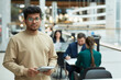 © Seventyfour - Waist up portrait of young man of Indian ethnicity holding planner and looking at camera in office building setting, copy space