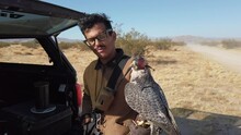 Medium Shot Of A Man Wearing Glasses Holding A Hooded Falcon Talks Into The Camera While Standing In The Arid Desert With Scrub Brush And Dusty Road
