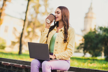 Poster - Portrait of beautiful trendy dreamy girl copywriter sitting on bench drinking latte using laptop traveling abroad outdoors