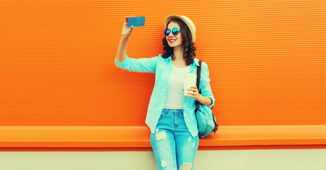 Portrait of happy smiling young woman taking selfie with smartphone with cup of coffee wearing summer straw hat, backpack on orange background, blank copy space for advertising text