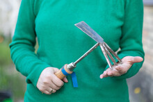 Caucasian Woman Gardener In The Garden Shows A Rake. The Concept Of Gardening And Sustainable Summer. Woman's Hand With A Garden Tool. Chopper Rake For Gardening In Female Hands. Place For Text.