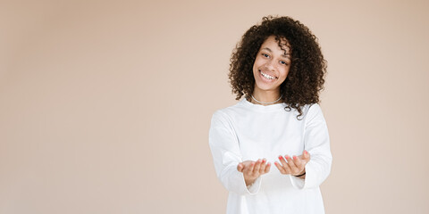 Portrait of smiling, pleasant African american girl studio portrait isolated on brown background showing something on the palms of her open two hands holding for object copy 