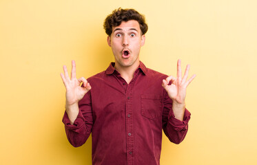 Wall Mural - young handsome man feeling shocked, amazed and surprised, showing approval making okay sign with both hands