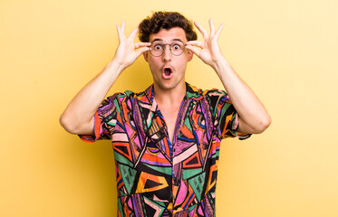 Wall Mural - young handsome man feeling shocked, amazed and surprised, holding glasses with astonished, disbelieving look