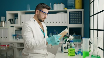 Sticker - Young hispanic man wearing scientist uniform reading book at laboratory