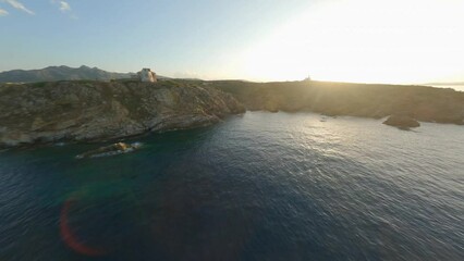 Poster - FPV video, view from above, stunning aerial view from an FPV drone flying at high speed over a rocky coastline with a lighthouse illuminated during a dramatic sunset. Faro di Capo Ferro, Sardinia.