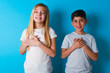 © Jihan - Honest two kids boy and girl standing over blue studio background keeps hands on chest, touched by compliment or makes promise, looks at camera with great pleasure.