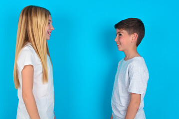 Wall Mural - Profile of smiling two kids boy and girl standing over blue studio background with healthy skin, has contemplative expression, ready to have outdoor walk.