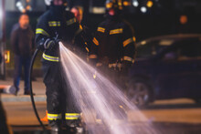 Group Of Fire Men In Uniform During Fire Fighting Operation In The Night City Streets, Firefighters With The Fire Engine Truck Fighting Vehicle In The Background, Emergency And Rescue