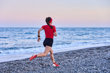 young athletic woman in a red shirt and braid running on the shore of the beach with mountains in the background