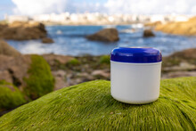 A White Cream Jar With A Blue Lid Stands On A Hill Of Green Smooth Grass. Close-up. The Grass Is Like Algae. In The Background Are Water And Stones. Naturalness. Environmental Compatibility. Spa. Body