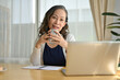 © bongkarn - A happy Asian aged woman sits at her home worktable in front of her laptop computer.