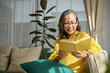 © bongkarn - Relaxed asian aged-woman in glasses sitting and reading a book in comfortable living room.