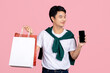 Smiling young handsome southeast Asian man holding mobile phone and shopping bags in pink studio isolated background