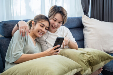 Wall Mural - Happy two lesbian woman, lgbtq homosexual Lesbian couples sharing earphones while listening music over smart phone in living room. Gay woman couple rest and relaxing by using smartphone in bedroom. 