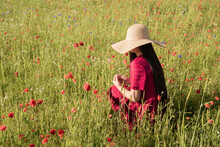 Woman With Summer Hat Sitting In Red Dress In A Field Of Poppies