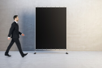 Poster - Side view of attractive young man walking past empty black rectangular poster in simple concrete interior. Advertisement and mock up concept.