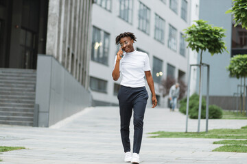 Cute African American girl in glasses standing on street and talking on cellphone on the background of the modern city. Portrait of joyful lady with dark curly hair in casual outfit standing on street