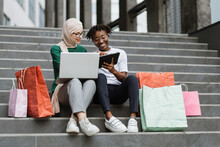 Two Pretty Multiracial Young Best Friends Women, Making Online Purchases Using A Tablet And Laptop Sitting On The Steps Of The Mall With Colorful Shopping Bags, Having Fun And Laughing Together.