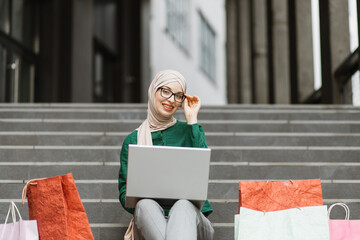 Wall Mural - Happy satisfied muslim girl, relaxing while sitting on the steps of a modern building rejoicing with laptop doing online order after shopping. Shopping in the city, urban lifestyle.