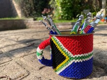 A Close Up View Of A Tin Cup With South African Beads Wrapped Around It With The South African Flag. There Are A Few Beaded Spoons On The Inside