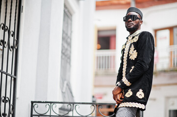 Wall Mural - Mega stylish african man in traditional jacket pose. Fashionable black guy in hat and sunglasses with cigar in hand.