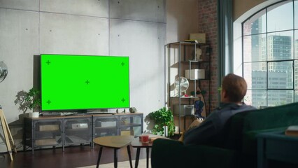 Aufkleber - In Loft Living Room an Attractive Man Sitting Down on a Couch, Turns On TV with Green Screen Mock Up Display with a Controller. Male Enjoys Leisure Time at Home.